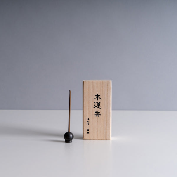 Kousaido wooden incense box with Japanese inscriptions, accompanied by a dark incense stick and a round black holder on a white surface with a gray background.