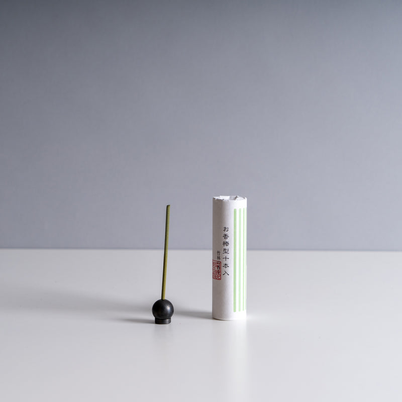 An upright incense stick with a dark round base stands next to a white cylindrical tube with green stripes and Chinese characters printed on it. Both are placed against a neutral gray background.