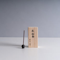 Wooden incense box with Japanese characters next to a dark incense stick held in a round black holder on a light gray background