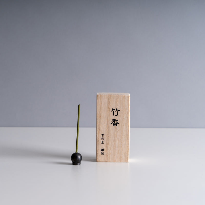 Kousaido wooden incense box with Japanese characters, accompanied by a green incense stick and a round black holder against a neutral backdrop.