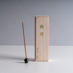 Light-toned wooden incense box with embossed Chinese characters, accompanied by a single brown incense stick and a small black holder against a neutral background.