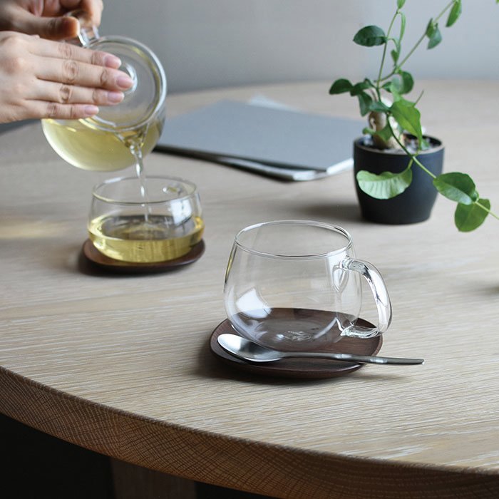 Two glass teacups on two Kinto 4" UNITEA Costers in Walnut. A person holding a glass teapot.